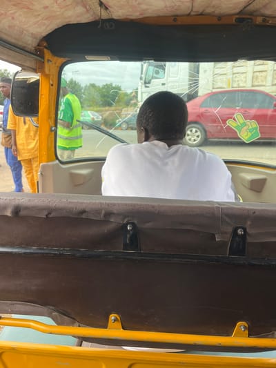 The image captures an interior view from a yellow tricycle, commonly known as a keke NAPEP, looking forward through a cracked windshield in Karmajiji, Nigeria. The central subject is a person, seen from behind, with short, dark, curly hair, wearing a plain white t-shirt, occupying the driver's seat.
The windshield is notably damaged, displaying a prominent crack originating from a central impact point, with numerous lines radiating outwards across the glass. Affixed to the upper right corner of the windshield is a vibrant green and yellow sticker depicting a hand making a peace sign (V-gesture).
Through the cracked and somewhat reflective glass, a bright daytime scene is visible. Elements outside include a red car, a white truck, and another person dressed in a bright green and yellow reflective vest, suggesting an urban or semi-urban setting. The interior of the tricycle features brown fabric covering structural pillars and parts of the roof, complemented by a lighter, patterned fabric on the ceiling. A side mirror is visible on the left, reflecting more of the exterior. The image appears to be a candid shot taken from a passenger's perspective during daylight hours.