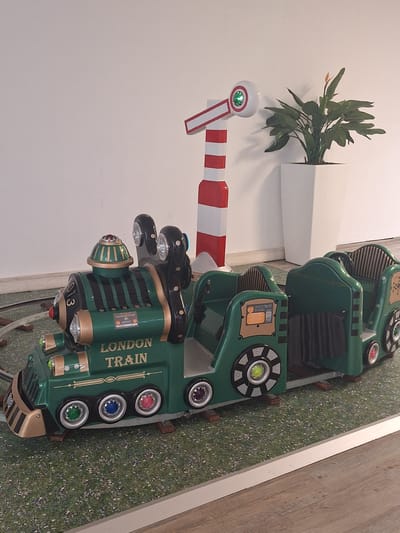 The image shows a green toy train labeled "LONDON TRAIN" with multiple passenger cars, positioned on a track. The train has decorative lights and a signal pole with red and white stripes next to it. A potted plant with green leaves and an orange flower stands behind the train. The scene is indoors, with a white wall in the background and a carpeted floor resembling grass. There are no people visible in the image. The image does not provide any information about the city of Bochum, Germany, or any specific location within it.