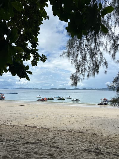 A serene beach scene unfolds in Pa Khlok, Thailand, under a cloudy sky. The foreground is dominated by sandy terrain with scattered debris and footprints. Beyond the beach lies the tranquil, blue ocean, dotted with numerous jet skis of various colors, suggesting a recreational area. In the shallow waters near the shore, a few people are visible, some standing on or near the jet skis, implying preparation for or the conclusion of water activities. The middle ground features a gentle swell on the water's surface. The background is comprised of a distant coastline with islands or peninsulas, appearing as hazy, green silhouettes against the sky. Lush green foliage, including branches and leaves of trees, frames the top portion of the image, adding a natural, slightly obscured perspective to the overall scene.
