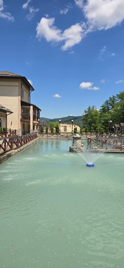 The image depicts a clear blue sky with scattered white clouds over a tranquil outdoor scene. In the foreground, a light turquoise-colored body of water features a fountain actively spraying water. A blue circular object floats in the water near the fountain. The water is bordered by a wooden fence with a stone base.

To the left of the water is a multi-story building with a beige facade and a dark-tiled roof. The building has balconies with wooden railings. Further to the right, a wooden bridge crosses the water, connecting to a stone structure that appears to be part of an older building or ruin. The background is characterized by lush green trees and rolling hills under the bright sky. The scene appears to be a resort or a recreational area. The location is identified as Babugent, Russia.