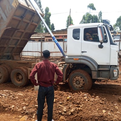 In Borabu Chitago ward, Kenya, a construction or excavation site is depicted under bright daylight. The main subjects are a white dump truck, a man, and a driver. The dump truck is positioned on muddy, reddish-brown earth, with its bed fully elevated by a prominent blue and silver hydraulic cylinder, suggesting it is either actively dumping material or has just finished. Its tires and lower body are heavily caked in mud, reflecting the challenging terrain.
In the foreground, a man is seen from behind, wearing a maroon long-sleeved shirt and dark jeans, with his hands on his hips, observing the truck's operation. His back is to the viewer, and his short, dark curly hair is visible. Inside the truck's cab, a person, presumably the driver, can be faintly seen wearing a striped shirt.
The background features several modest buildings with corrugated metal roofs and walls, typical of a rural or semi-urban setting. A corrugated metal fence runs across the mid-ground. Tall, slender trees are scattered throughout the background, indicating an open environment.
Visible text includes "PER" on the hydraulic cylinder. A sign in the far right background displays "NYAMAIY CV/CHEMIS WA HAPA HAI". Through the truck's passenger window, faint text "Co-op bank" and "safaricom" are discernible on posters within a distant building. The sky appears bright and possibly hazy or overcast, characteristic of daytime.