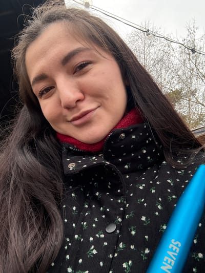 The scene is an outdoor setting in the city of Kurovskoye, Russia. A female subject is captured in a close-up selfie, facing the camera with a slight smile. She has long, dark brown hair and is dressed in a black jacket featuring a white and green floral pattern, worn over a red, textured collar. A bright blue cylindrical object, bearing the white text 'SEVEN,' is partially visible on the lower right side of the frame. The background consists of an overcast sky, leafless trees, and a string of clear globe lights, suggesting a temperate, possibly late-season, environment.