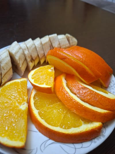 The image shows a plate of sliced bananas and oranges on a dark wooden surface. The bananas are sliced into short, thick pieces and arranged in a row on the left side of the plate. The oranges are sliced into wedges and are layered on top of each other on the right side of the plate. The plate is white with a subtle floral pattern. The lighting is natural and highlights the textures of the fruit. There are no people or animals visible in the image. The scene appears to be indoors, possibly in a kitchen or dining area. The location in Vagharshapat, Armenia, is not determinable from the visual information provided in the image.