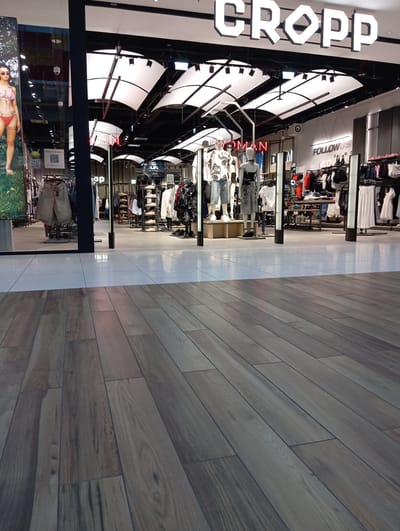The image shows the exterior of a CROPP clothing store in the city of Wołomin, Poland, located in a commercial area or mall. The storefront features a large, illuminated "CROPP" logo. Inside, mannequins are displaying clothing, and racks of garments are visible, suggesting a retail environment. The floor in the foreground is made of wood-like tiles, and the area behind it is tiled with lighter-colored tiles. There are no identifiable people in the visible portion of the scene.