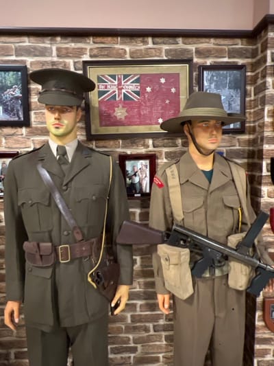 Two mannequins dressed in military uniforms are displayed against a brick wall adorned with framed images, including one of the Australian flag. The upper mannequin wears a brimmed hat and carries a firearm. The lower mannequin also wears a military hat and is equipped with a belt and holster. The scene is likely part of a museum or historical exhibit.