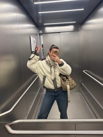 An individual is depicted taking a mirror selfie inside a brushed stainless steel elevator located in Tourcoing, France. The subject, a person with dark hair pulled back, is wearing a cream-colored fleece jacket with olive-green trim, blue denim jeans, and a light-colored patterned scarf. A patterned shoulder bag with a light beige strap hangs from their left shoulder. The subject holds a light-colored smartphone in their right hand, obscuring their face, while making a two-finger 'V' gesture with their raised left hand, which also holds a set of keys with multiple keyrings and possibly spectacles. The elevator features a dimpled metal floor, horizontal stainless steel handrails, and linear LED lighting on the ceiling. A vertical panel labeled "EE A" is visible on the left wall.