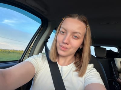 A woman is taking a selfie in a car. She has blonde hair and is wearing a white t-shirt and a seatbelt. The car is on a road with trees and fields visible outside the window. The sky is blue with some clouds. The image is taken from a slightly low angle.