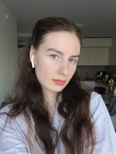 An adult female, with long dark brown hair and blue eyes, is depicted from the upper chest, facing forward with a neutral expression. She wears a light purple and white striped collared shirt and has a white earbud inserted into her left ear. Her lips show a light pink tint. The background is an indoor residential setting, featuring a modern kitchen or kitchenette area. Visible elements include light-colored upper cabinets, a sink with a faucet, and a countertop displaying various kitchen appliances such as a stand mixer and a coffee maker. A vase with white flowers is present on the counter to the right. A grey blanket or throw is partially visible in the lower right background. The scene is located in Ковалёво, Russia.