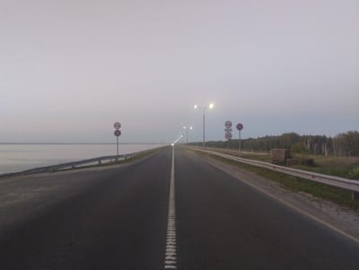 The image shows a straight, paved road in the city of Zavolzhye, Russia, likely at dusk or dawn due to the ambient light. The road has a white dashed line down the center and extends into the distance where it is illuminated by streetlights. On the left side of the road, there is a body of water, possibly a river or lake, with a metal guardrail along the edge. On the right side of the road, there is a grassy area with trees in the background and more guardrails. Several traffic signs are visible: a "no entry" sign and a speed limit sign indicating 50 km/h on the right side, and two "no entry" signs on the left. There are no people or vehicles visible in the image. The sky is overcast and light grey.