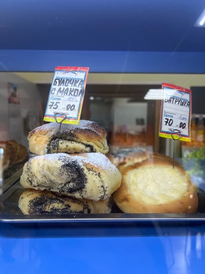 This media file shows a close-up view of baked goods displayed in a refrigerated glass case, likely in a bakery or food store in Lomonosov, Russia. The primary subjects are two types of pastries: a large, round pastry with a creamy filling (likely cheese, resembling a Vatrushka), and several smaller, twisted pastries covered in poppy seeds and powdered sugar.

Attached to each type of pastry is a small, colorful price tag. The tag for the Vatrushka, with red and yellow accents, indicates "ВАТРУШКА" (Vatrushka) as the name, "Лит" (Lit) as the manufacturer, and a price of "70.00". The tag for the poppy seed pastries, with blue and yellow accents, reads "БУЛОЧКА С МАКОМ" (Bun with poppy seeds), lists the weight as "100г" (100g), and a price of "75.00".

The setting is indoors, with the baked goods presented in a clean, professional display. The background is blurred but suggests the interior of a commercial establishment, possibly with other food items or shelving visible. The lighting is bright, highlighting the textures and colors of the pastries. There are no people visible in the frame. The overall impression is one of fresh, appetizing baked goods ready for purchase.