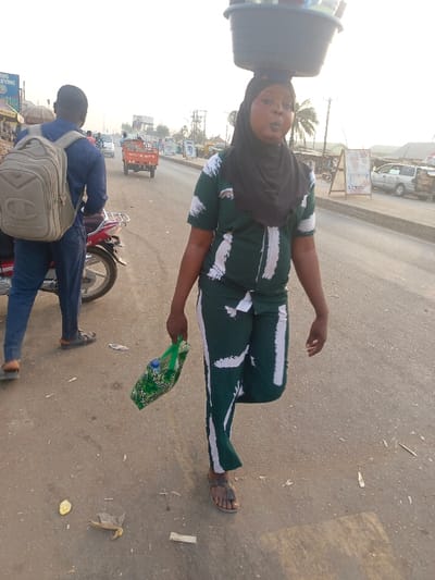 This image depicts a vibrant street scene in Alizaga, Nigeria, under what appears to be bright, hazy natural light, possibly indicating late afternoon or early morning.
In the foreground, a woman walks towards the right side of the frame, balancing a large, dark basin or bowl on her head, characteristic of street vendors. She is dressed in a dark green two-piece outfit featuring white abstract brushstroke patterns and wears a black headscarf. In her left hand, she carries a green bag containing a plastic bottle. Her mouth is slightly open, suggesting she might be speaking or calling out to potential customers. She is wearing simple sandals.
To the left and slightly behind her, a man stands with his back mostly to the camera. He wears a blue long-sleeved shirt, blue trousers, and sandals. A light brown backpack with a prominent "Champion" logo is visible on his back. He is positioned next to a red motorcycle or scooter, only partially visible.
The setting is an asphalt road with some litter scattered on the ground. In the background, a red three-wheeled vehicle, possibly a cargo tricycle, is visible on the road, alongside other indistinct vehicles and roadside structures. Faint text on a sign behind the man might read "WING BUILDING," and other distant signs are illegible. The scene captures a moment of daily life and commerce on a local thoroughfare.