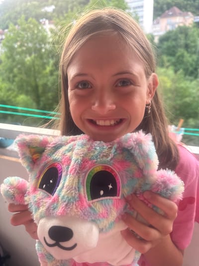 This is a close-up, tilted, outdoor portrait of a young girl holding a colorful plush toy. The girl has long, light brown hair, blue eyes, and is smiling with her mouth open, revealing her teeth. She is wearing a pink t-shirt. The plush toy is rainbow-colored, with large iridescent eyes that have stars in them. One of the girl's hands is holding the toy. The background is blurred, showing green trees and a building, suggesting an outdoor setting. The lighting appears natural, indicating it is daytime, and the weather seems pleasant, given the outdoor scene. The overall emotion conveyed is joy and playfulness. No text is visible in the image.