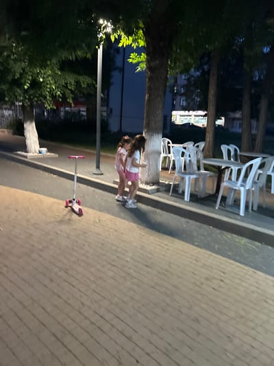This image captures an outdoor urban scene at night in Dzhebel, Bulgaria, illuminated by artificial light. The foreground features a light-colored paved surface, possibly interlocking bricks or pavers, laid in a diagonal pattern. To the left, a children's three-wheeled scooter, predominantly white with pink accents on its wheels and handlebars, rests upright on this paved area.
Mid-ground, two young girls are standing near the base of a tree. The girl on the left wears a light pink top and shorts, while the girl on the right wears a white top and pink shorts and appears to be holding a small, light-colored object in her left hand. Both have long, dark hair.
A row of mature trees with dark trunks and foliage runs along the right side of the image. The base of at least two tree trunks is painted white. Beneath these trees, an outdoor seating area is visible, comprising multiple white plastic chairs arranged around at least two round tables with dark bases.
A tall street lamp with a bright light fixture is positioned towards the center-left, providing the primary illumination for the scene, casting a yellowish glow on the tree foliage and paved ground. In the background, dimly lit buildings with windows are visible through the trees, one displaying a bright, possibly illuminated, sign or interior light. A darker asphalt surface, likely a road or path, is present beyond the paved area and curb. A small white bucket or container is situated at the base of a tree on the left.