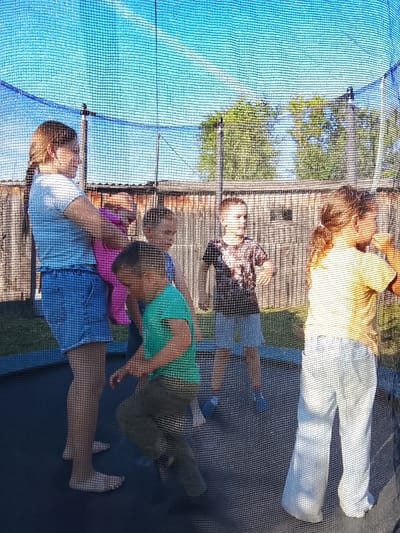 An outdoor image taken from within a trampoline enclosure shows five children of varying ages on the trampoline, with a wooden fence and trees in the background under a clear blue sky. The trampoline is enclosed by a black safety net. On the left, a girl with braided hair in a light blue t-shirt and denim shorts stands holding a toddler wearing a pink outfit. To her right, a boy in a green t-shirt and dark pants is mid-jump, with his legs extended. Further right, another boy in a dark patterned t-shirt and light blue shorts is facing left, with his arms slightly raised. On the far right, a girl with blonde hair in a yellow t-shirt and light blue pants is facing the other children, her hands raised near her face. The scene appears to be a residential backyard in the city of Kuyeda, Russia.