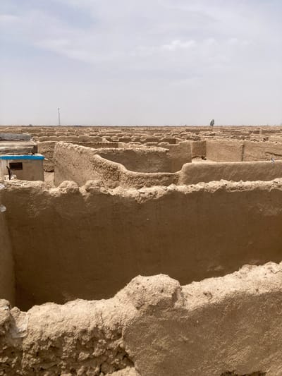 The image depicts a vast, arid landscape dominated by an extensive array of ancient or traditional mud-brick structures, likely kashans or a settlement, stretching into the distant horizon under an overcast sky. The photograph, correctly oriented, shows these earth-colored formations filling the right side of the frame, extending from the foreground to the background.

The structures are characterized by their multi-layered, stepped, and heavily eroded appearance, suggesting significant age and exposure to weather. They are constructed from what appears to be sun-dried mud or adobe, with visible textures of coarse earth and embedded fibers (possibly straw) in the close-up views. The edges of these formations are irregular and jagged due to extensive weathering.

The sky occupies the left half of the frame and is a uniform, light gray, indicating overcast conditions or a hazy atmosphere. The diffused light results in soft shadows and a general lack of harsh contrasts across the scene.

In the foreground, towards the right, a small, partially obscured structure with a distinct blue-painted section and a small, dark rectangular opening is visible, offering a subtle hint of recent human presence or habitation amidst the older ruins. Further in the distance, a solitary, sparse tree stands out against the muted backdrop. A faint, slender white pole or tower is also barely discernible on the distant horizon.

No human activity or individuals are visible in the scene. The overall impression is one of stillness, desolation, and historical depth. Given the context of Saranan, Pakistan, these structures are indicative of traditional architecture adapted to a desert or semi-desert environment, reflecting a rich cultural heritage. There is no visible text in the image.