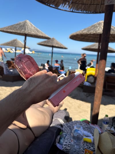 A close-up shot shows a person's hand holding a pink water gun on a sunny beach. The person's arm and leg are visible in the foreground, with the arm hairy and the leg smooth, suggesting a man's arm and a woman's leg. The water gun is white and pink and has text that reads "A CASE COLLECTION" and "WATER GUN SHARK MK." In the background, beach umbrellas, lounge chairs, and people can be seen, as well as the sea. The overall scene depicts a relaxed day at the beach in Nea Potidea, Greece.