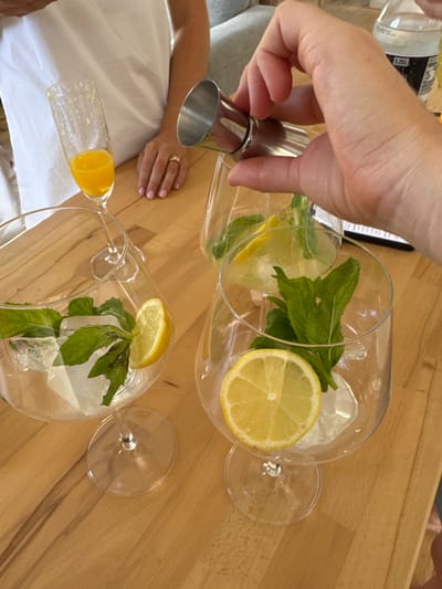 Two glasses of mojitos are being prepared on a wooden surface. One glass is nearly full, containing a lemon slice, mint leaves, and ice cubes. The other glass is partially filled with a yellow liquid, possibly juice or syrup. A hand is pouring liquid from a stainless steel measuring cup into the glass, suggesting the addition of another ingredient. The scene is brightly lit and suggests a domestic setting.