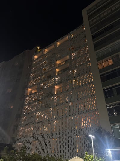 The image shows the facade of a building in Rio de Janeiro, Brazil at night. The building features a distinctive white, perforated concrete screen with an intricate, repeating pattern of oval shapes. Through these openings, warm yellow light emanates from within the building, illuminating the facade. Some windows and balconies are visible behind the screen. To the right of this building, another taller, darker building with a green facade and numerous windows is partially visible. The sky above is dark, indicating nighttime. In the foreground, lush green foliage and a modern street lamp are present. The overall impression is one of modern architecture and urban nighttime ambiance.