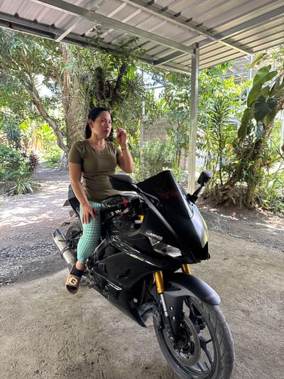 The scene, set in Carmen, Philippines, features an adult female seated on a black sport motorcycle beneath a grey corrugated metal roof supported by metal beams. The woman, with dark hair tied back, wears an olive green t-shirt, blue and yellow checkered leggings, and brown slide sandals with a white 'G' emblem. She is positioned with her left foot on the concrete ground, her right leg bent against the motorcycle, her left hand resting on her right thigh, and her right hand raised to her mouth area. Her gaze is directed slightly upwards and to her right. The motorcycle is black, featuring gold front suspension forks, a dark tinted windshield, and a 'LUMINOS LED' decal on the fairing. A 'YAMAHA' logo is present on the front fender, and a silver exhaust pipe extends from the left side. The immediate ground is concrete, transitioning to a gravel path to the left and rear. The background is dominated by lush tropical foliage, including trees and various green plants, under bright daylight.