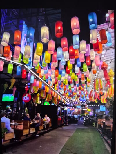 The image shows a night scene with colorful lanterns suspended above an outdoor seating area. The lanterns are cylindrical and in a variety of colors. Below the lanterns is a seating area with wooden tables and chairs. People are sitting at some of the tables, appearing to be enjoying themselves. In the background, there are buildings with various signs and lights, as well as a television screen. The image does not seem to be in Mont-Saint-Martin, France.