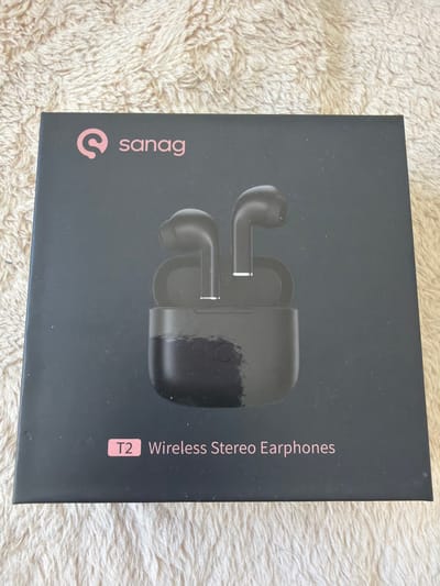 The image shows the front of a black product box for Sanag T2 Wireless Stereo Earphones. The box is placed on a light-colored, textured surface, possibly a rug or carpet. The box has the Sanag logo and name printed in pink on the left side. To the right of the logo, there is a graphic illustration of the wireless earphones and their charging case. Further to the right, the text "T2 Wireless Stereo Earphones" is printed vertically in pink. The overall impression is that of a product advertisement or packaging. There are no people visible in the image. The lighting appears to be indoor lighting, casting a soft illumination on the box. No specific time of day or weather conditions can be inferred. The location context of Schwedt/Oder, Germany, does not provide any direct visual cues within the image itself.