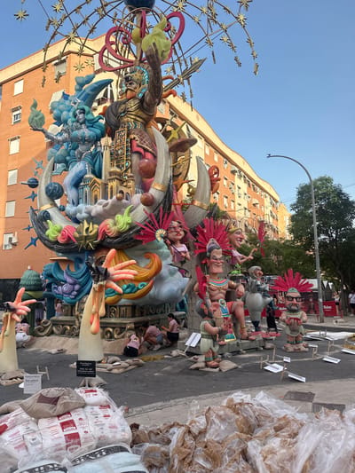 In Alicante, Spain, a large, colorful artistic installation dominates the scene, featuring several larger-than-life figures. The central element is a towering structure adorned with mythological or fantastical characters, including a warrior in golden armor, a blue-skinned female figure with flowing hair, and other cartoonish characters with exaggerated features and vibrant headdresses. The installation is intricately detailed with elements like a crescent moon, planets, and ornate architectural motifs.

The figures are placed on a street or plaza, with a backdrop of a multi-story brick building. Piles of materials, including sacks and plastic-wrapped objects, are visible in the foreground, suggesting ongoing construction or preparation. Several people are present, some appearing to be working on the installation, while others observe from a distance. The sky is clear and blue, indicating daytime. The overall impression is of a public display, likely part of a festival or event.
