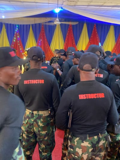 The image displays an indoor gathering in Kachia, Nigeria, featuring a dense congregation of individuals. Multiple people, primarily male, are visible from behind or in profile in the foreground and midground. They are uniformly dressed in black long-sleeved shirts with "INSTRUCTOR" in red capital letters printed across the upper back, camouflage pattern trousers, and black baseball caps. Several individuals have walkie-talkies clipped to their belts. The event takes place in a decorated venue, likely a hall or large tent, characterized by white and blue fabric drapes on the ceiling. The rear wall is adorned with alternating vertical fabric panels in blue, yellow, and red, with string lights embedded within the red panels. Overhead lighting and a ceiling fan are visible. The floor is covered with a red carpet. Participants are standing; some appear to be engaged in conversation or utilizing mobile phones, with one person in the background holding a phone vertically.