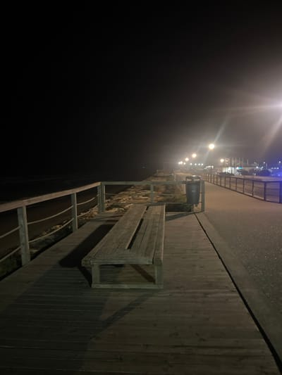 An empty wooden bench sits alongside a dark, narrow pathway. A waist-high metal railing edges the path. Above, streetlights illuminate a sidewalk, creating stark highlights on the weathered wood. A trash receptacle is positioned near the bench. The scene evokes a lonely, possibly foggy night.