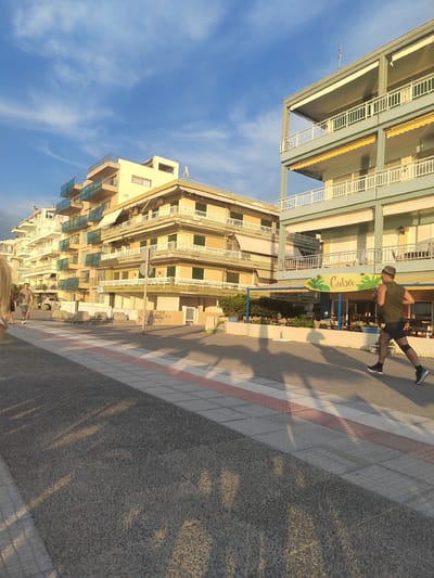 In Perea, Greece, on a paved promenade, a man is running towards the right of the frame. He is wearing a dark sleeveless shirt, black shorts, a headband, and black and white athletic shoes. The promenade is bordered by a sidewalk featuring a pattern of grey tiles with a red and white stripe running through it, and a dark asphalt surface. In the background, a multi-story building with light yellow and light blue facades features numerous balconies with white railings and blue glass. A surfboard-shaped sign with the word "Cuba" and palm tree graphics is mounted on the building's lower level, above what appears to be an outdoor seating area. To the left of the running man, other people are visible walking, and there are several concrete bollards and trees. The sky is a clear blue with scattered white clouds, and the scene is illuminated by the warm light of what appears to be late afternoon or early evening sun, casting long shadows.