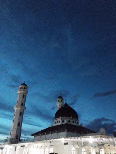 In the city of Barat Yaman, Indonesia, a mosque stands against a deep blue twilight sky filled with wispy clouds. The mosque features a prominent dark dome and a tall, slender white minaret with arched windows. The building's facade is visible, with a covered walkway featuring arched openings and exterior lighting. The image captures the mosque from a low angle, emphasizing its architectural grandeur against the dramatic evening sky. No people are clearly visible in the frame.