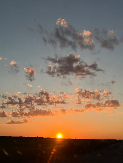 This is a vertical, eye-level, medium shot photograph of a sunset over a rural landscape, presumably near Skomorokhy, Ukraine. The sky dominates the upper two-thirds of the frame, transitioning from a pale blue at the top to a vibrant orange near the horizon. Scattered clouds are present throughout the sky. Some clouds are dark gray and appear heavy, while others are illuminated by the setting sun, glowing with shades of pink and orange. The sun itself is a bright, orange orb low on the horizon, casting a golden light across the scene and creating a lens flare effect in the lower portion of the image. A dark, silhouetted treeline forms the horizon line, separating the sky from the land. The foreground consists of dark, out-of-focus land, likely fields, and a dirt path on the right side of the frame. No people or animals are visible in the image. The overall mood is serene and peaceful, characteristic of a sunset.