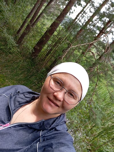 A woman wearing a white head covering and glasses is taking a selfie outdoors in a forest. She is wearing a dark blue hooded jacket with a zipper. The background is filled with trees and greenery. The location is Vekoshinka, Russia.