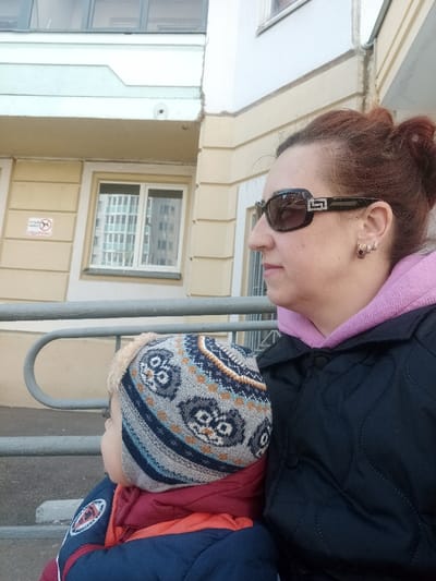 An adult female and a young child are depicted outdoors in the city of Serpukhov, Russia. The adult female, with reddish-brown hair tied back, is viewed in profile facing right. She wears dark sunglasses with prominent silver-colored, possibly rhinestone-encrusted, Greek key pattern temples, a light pink hooded sweatshirt, and a dark, possibly quilted, jacket. Small earrings with reddish stones are visible on her right ear. To her left, partially obscured, is a young child viewed in profile facing left. The child wears a multi-colored knitted winter hat with a patterned design (possibly owls) and faux fur trim, along with a red and blue winter jacket. A light-colored metal handrail extends horizontally across the middle ground. Behind the subjects, a multi-story building with light yellow walls and white trim is visible. A white, rectangular sign with a red "no dogs" symbol and Cyrillic text ("ВЫГУЛ СОБАК ЗАПРЕЩЁН" - Dog walking is prohibited) is affixed to the building's facade. A white-framed window on the building reflects a grid pattern from another structure. The scene is illuminated by natural light.
