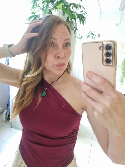 A woman is taking a mirror selfie in what appears to be a restroom. She has light brown hair, green eyes, and fair skin with freckles. She is wearing a maroon, one-shoulder top with ruching on the side and a necklace with a green pendant. On her left wrist, she is wearing a light gray smartwatch. She is holding a light pink smartphone in her right hand, which is angled to capture the reflection in the mirror. The background shows a white tiled floor, a white wall, and several green plants in the upper portion of the image. There are also some vertical blinds or a door visible on the left side. The lighting is bright, suggesting an indoor setting with natural light. The image does not provide any specific details that would indicate the location is Kemul, Russia.