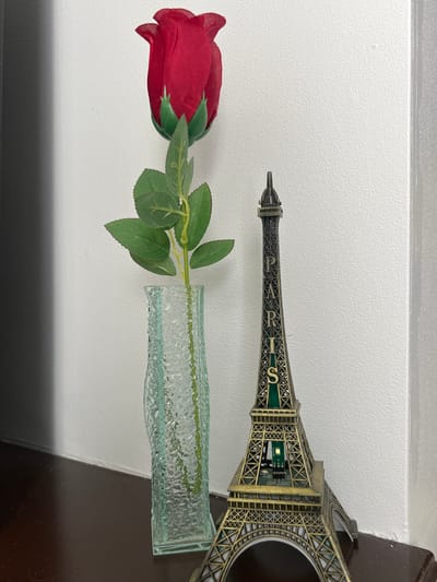A red artificial rose in a clear, textured glass vase lies next to a miniature Eiffel Tower souvenir on a surface. The surface appears to be adjacent to a wall, which is white. The darker, possibly wooden, surface suggests the display is inside, possibly on a shelf or table. The Eiffel Tower souvenir has "PARIS" inscribed on it. The scene does not provide information about Gorleston-on-Sea, United Kingdom.