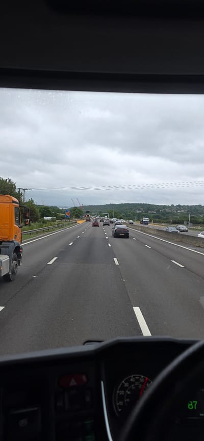 The image is taken from the interior of a vehicle, likely a truck, looking out through the windshield at a highway. The sky is overcast with clouds. In the foreground, the dashboard of the vehicle is visible, including a red hazard light button, a speedometer showing a speed of 87, and a portion of the steering wheel. The highway is multi-lane, with traffic including cars and trucks. There are white road markings and a concrete barrier separating lanes and the side of the road. On the left side of the highway, there is an orange truck. In the distance, there are trees and hills, with some construction cranes visible. The location is the M1 motorway in Sheffield, United Kingdom, heading north.