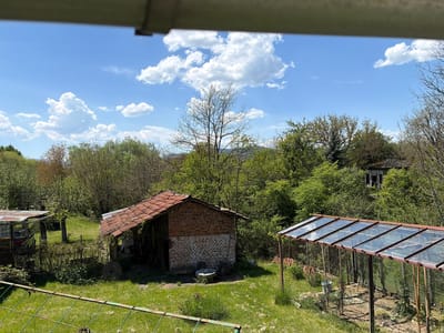 The scene is a view of a rural landscape, likely in Ravnishte, Bulgaria, as indicated. The foreground includes a grassy area with a low, green metal fence. To the left, there's a small shed with a red tiled roof and brick construction. A greenhouse is visible to the right, its frame constructed of metal, with what appears to be glass or plastic paneling. The greenhouse stands on bare ground. In the middle ground, there are various trees and dense foliage, partially obscuring a building in the background. The sky is blue with scattered white clouds. The image is taken from an elevated position, looking down upon the scene. A structure is visible at the top of the frame, possibly part of the building from which the image was taken.