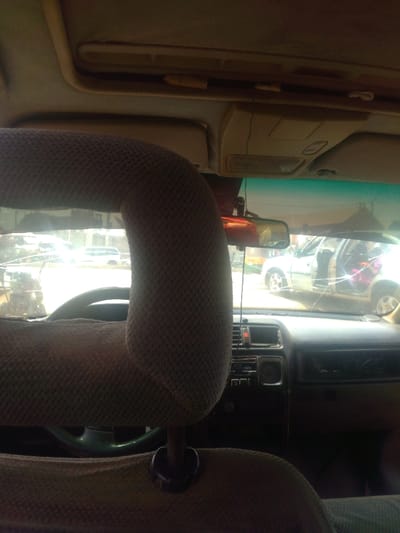 The image presents a view from inside a vehicle, looking forward through a cracked windshield onto an outdoor scene in Mista Ali, Nigeria. Dominating the immediate foreground is a textured, light brown fabric headrest, likely from the driver's seat. Partially visible below and to its left is a dark steering wheel. The vehicle's dark-colored dashboard, equipped with controls and vents, extends across the lower portion of the frame behind the headrest. The light-colored overhead headliner of the vehicle features compartments and a yellow, cylindrical object tucked into a trim piece. A rearview mirror hangs centrally from the headliner. The windshield exhibits significant cracking, particularly on the right side, and has a blue-green tint at its top edge. Through the fractured windshield, under bright daylight, another light-colored vehicle, possibly silver or white, is visible in the midground with its front passenger door open, revealing a pink object inside. Additional indistinct vehicles or structures occupy the background. No individuals are visible.