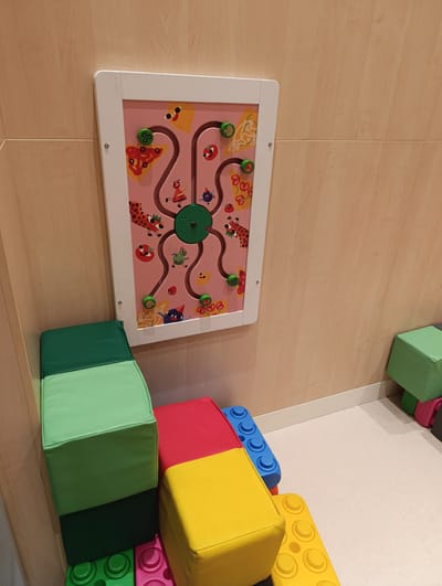 This image shows a wall-mounted bead maze toy with colorful cartoon characters and food items on a pink background. The maze has green beads that can be moved along the paths. The toy is mounted on a light-colored wooden wall. Below the toy, there are large, soft, block-shaped cushions in green, red, and yellow, resembling oversized toy building blocks. A green cushion is stacked on top of a dark green cushion. In the foreground, the yellow cushion is placed next to a blue block, and part of a pink block is visible. To the right, a small green cube is placed on the floor. The setting appears to be an indoor play area or a child's room, with a light-colored carpet. There are no people visible in the image, and the lighting suggests it is daytime. The image is taken from a slightly elevated angle, looking down at the cushions. There is no visible text in the image.