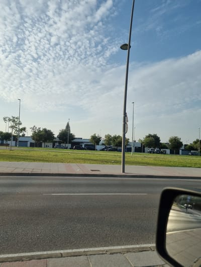 The image shows a roadside scene in Jerez, Spain. The perspective is from inside a vehicle, looking out. A side mirror is visible in the upper right corner, reflecting the sky and road. The main scene depicts a street with a sidewalk and a grassy verge lined with trees. Beyond the verge, there are modern buildings, possibly apartments or offices, with parked cars visible. A distinctive, tall, conical structure resembling a Christmas tree or a stylized sculpture rises above the trees in the middle distance. Streetlights are positioned along the road. The sky is blue with scattered white clouds, suggesting it is daytime. The overall impression is one of a modern urban or suburban environment on a clear day. No people are clearly visible in the image. No text is discernible.