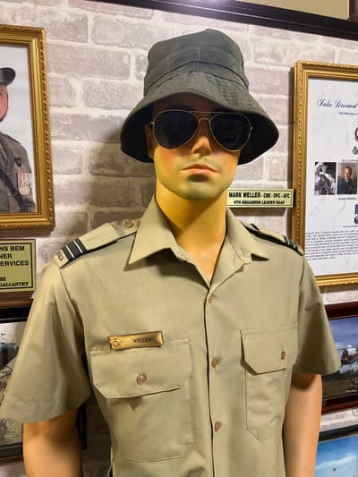 A mannequin, positioned at an angle, sports a khaki uniform with a "WELLER" name tag and epaulettes. The mannequin's head is adorned with a dark green bucket hat and aviator sunglasses. "Australia" is visible on its shoulder.
Behind the mannequin is a faux-brick wall adorned with several framed items, including information placards. One placard identifies "MARK WELLER CBE-DFC AFC, 9TH SQUADRON LEADER RAAF." Another reads "John Desmond, 1st February." A plaque below features "NS BEM" and "GALLANTRY."