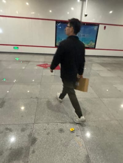 A person wearing dark clothing and white sneakers walks on a tile floor. They are carrying a brown package. A display screen with an image on it is to their left. A red arrow pointing left is on the floor. There is a small, yellow object on the floor. Green exit signs are visible on a wall to the person's left.