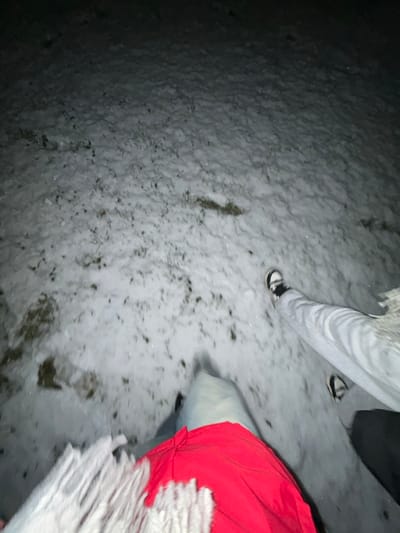 An uneven surface covered in snow and patchy dead grass occupies most of the frame. Footprints are visible in the snow. Two individuals are partially visible, one wearing light grey sweatpants and black and white sneakers, the other in blue jeans and a bright red jacket with a white furry collar. The scene is dimly lit, with the left side appearing darker than the right.