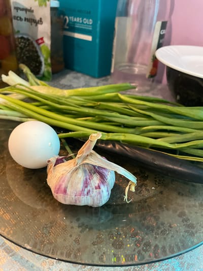 A high-angle, tilted close-up shot depicts a glass plate containing an egg, a head of garlic, scallions, and a dark-colored, unidentifiable object. The scene appears to be indoors, with items stacked haphazardly in the background. The garlic has purple and white stripes on its outer layer, and the scallions are green and white. The egg is white and round. The photo provides no information to establish that this photo was taken in Olginka, Russia.