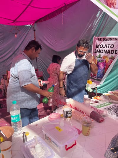 The scene is a food stall in a market in Chandigarh, India, at the coordinates 30.764, 76.798. There are three people in the scene. One man, wearing a plaid shirt, is holding a green bottle and pouring liquid into a container. Another man, wearing a white shirt and a blue apron, is wiping his face with a napkin. A woman in a pink shirt is standing in the background. The stall has a pink and white canopy, a table with various food items, containers, and beverages. The table has a sign that reads "Mojito Lemonade," and there are signs with a QR code and the words "Fruit Flavours." There are empty cups, a bottle of water, and a bottle of soda on the table. The scene appears to be a busy street market in Chandigarh, India.