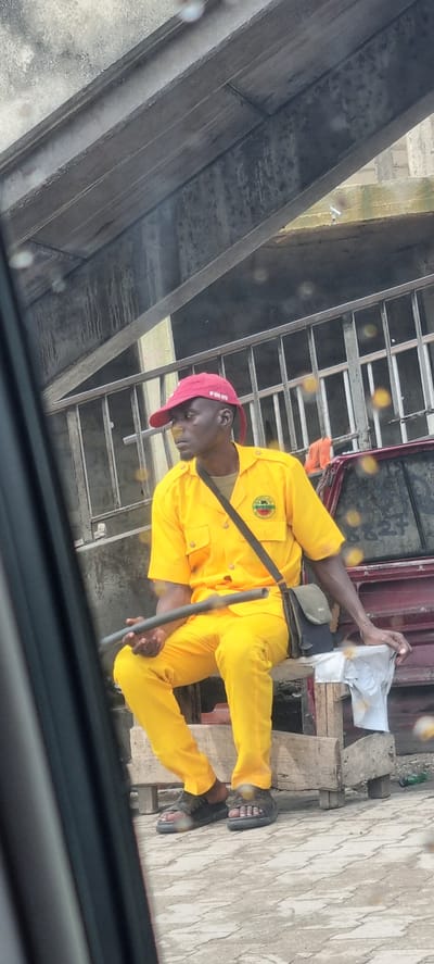 A man wearing a bright yellow uniform consisting of a short-sleeved shirt and pants sits on a wooden structure. He has a red baseball cap on his head, and a dark-colored bag is slung across his chest. In his hands, he holds a dark, cylindrical object. The man appears to be in a resting or waiting posture. The background suggests an urban environment, possibly under an overpass or bridge, with a metal railing visible behind him. The ground is paved with interlocking bricks. The scene is located in Iganmu, Nigeria.