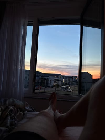 The image shows a first-person perspective from a bed looking out a window at dusk. The window frames a view of a residential area with multi-story buildings and a street. The sky displays a gradient of colors from blue at the top to orange and pink near the horizon, indicating sunset. There are several vehicles visible on the street, including a red van and a white camper van. A white sheer curtain is drawn on the left side of the window. The foreground of the image shows a person's legs and feet resting on a bed with blankets. The scene appears to be set in the city of Bönnigheim, Germany, given the architectural style of the buildings.