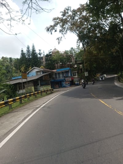 The image displays a daytime scene in Khajuripada, India, featuring a two-lane asphalt road that curves uphill to the right. On the immediate left, a black and yellow striped metal guardrail borders a concrete sidewalk, alongside a solid white line marking the road's edge. Further right on the road, a faded yellow broken line is visible.
A cluster of buildings is situated on a slope to the left of the road. In the mid-ground, a multi-story structure with a blue lower facade houses multiple shopfronts, exhibiting various signage and products. Above this, another building with white walls and a light blue roof is visible. Elevated in the background, a prominent building with a large dome on its roof stands amidst dense green foliage, including tall pine-like trees. The text "HO ZAR" is discernible on this building's facade.
On the road, at least five motorcycles are traveling away from the viewer in the right lane, followed by a white van further down the curve. Dense green vegetation and trees line both sides of the road and cover the surrounding hillsides. Thin power lines are visible stretching horizontally across the light grey, partly cloudy sky.