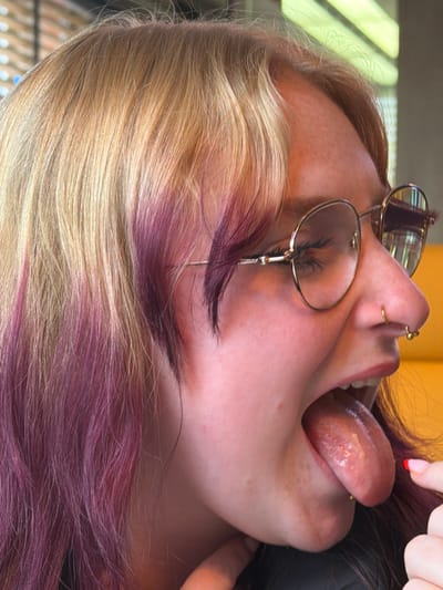 The image shows a close-up of a person's face with their head tilted back and mouth open, tongue extended. The person has fair skin, freckles, and blonde hair with purple streaks. They are wearing glasses with gold frames and have piercings in their nose and tongue. A hand with a painted fingernail is visible near the top right corner of the image. There is no information about the location in the city of Grevenmacher, Luxembourg.