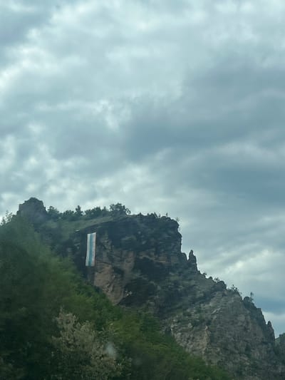 The image shows a view of the rocks and vegetation around Bukovo, Bulgaria. A horizontally oriented rectangle with three stripes, white on top, light blue in the middle, and blue at the bottom, is attached to the rockface. The sky is cloudy.